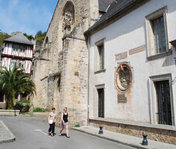 Cette photo offre une vue d'une partie de la façade du site des Jacobins. Au premier plan, sur la droite, se trouve une partie du bâtiment conventuel (c'est à dire du couvent). Deux femmes se dirigent vers l'entrée du Musée en marchant. Dans le prolongement de cette façade, se dresse l'église des Jacobins. Tout au fond de cette image, une jolie maison à pans de bois rouges, perpendiculaire à l'église laisse deviner le passage de la rue des Vignes qui longe le site des Jacobins.