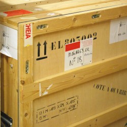 cette image réalisée pour illustrer le transport des œuvres est une photographie d'une caisse en bois spécialement dédiée aux déplacement des objets fragiles. Sur les trois faces de cette caisse, des lettres, des étiquettes attestent de nombreux déplacements déjà réalisés.