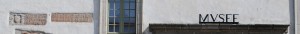 Cinq lettres noires de style antique sont posées au dessus d'une longue pierre qui semble être le haut d'un porche. Ces lettres indiquent l'entrée du Musée de Morlaix. Elles écrivent MUSEE. Sur la moitié gauche de ce bandeau une fenêtre et des pierres comportant des inscriptions ressortent de la pâleur de ce mur du bâtiment des Jacobins.