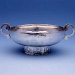 Sur un fond bleu, est posée une coupe de mariage en argent datant du milieu du 18ème siècle. Le bol de cette coupe à deux anses, posé sur un piédouche décoré, est gravé O. Peront.