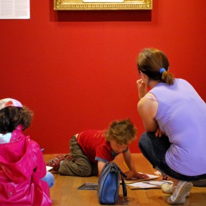A droite de cette image, une femme est accroupie, de dos. Elle porte un petit haut violet et un jean. Elle aide son fils qui travaille installé par terre dans la salle d'exposition des Jacobins. A gauche, une petite fille assise, de dos qui porte un manteau rose est en partie visible. Les trois personnes sont face à un mur rouge sur lequel la baguette dorée du bas d'un tableau apparait.